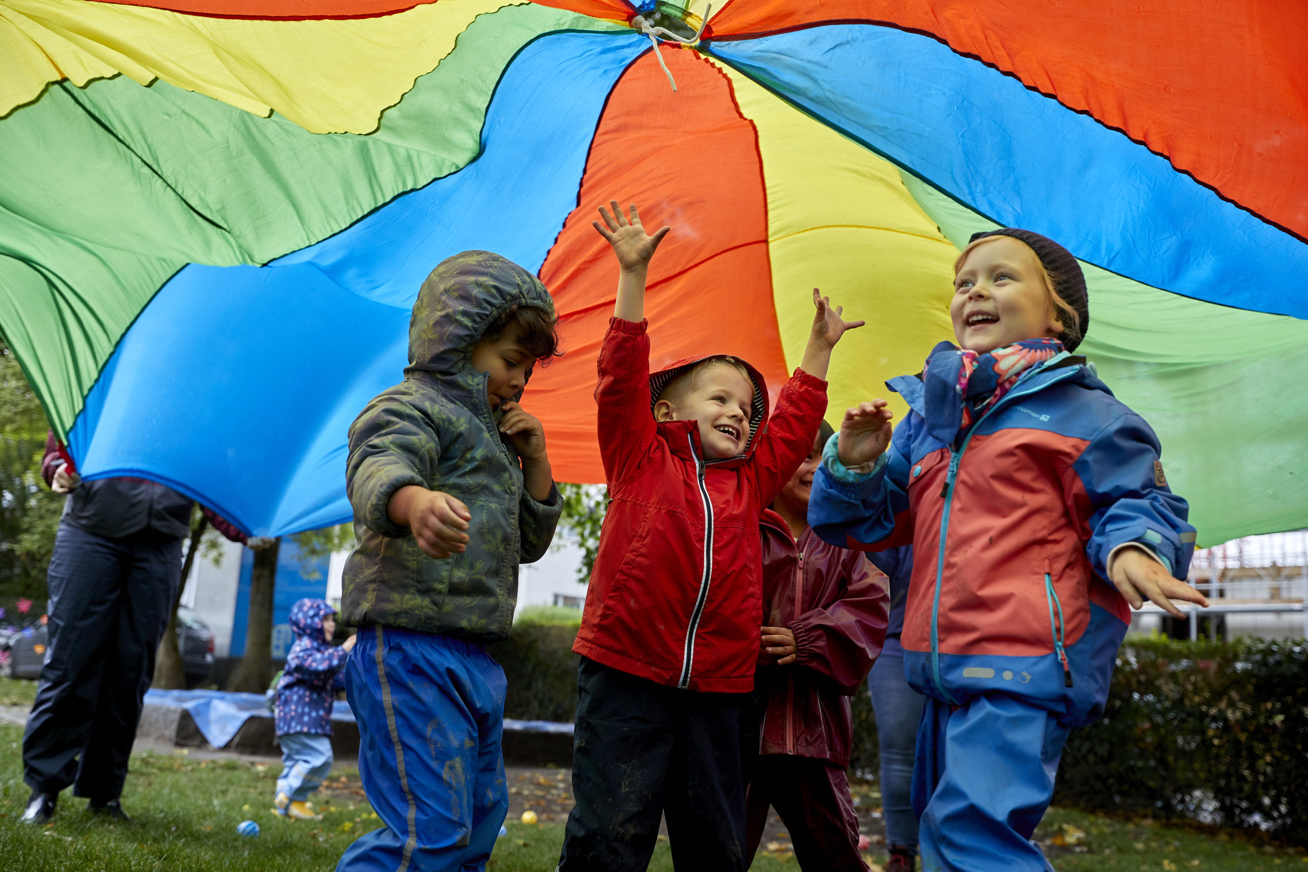Kinder spielen in Regenkleidern unter dem Regenbogentuch
